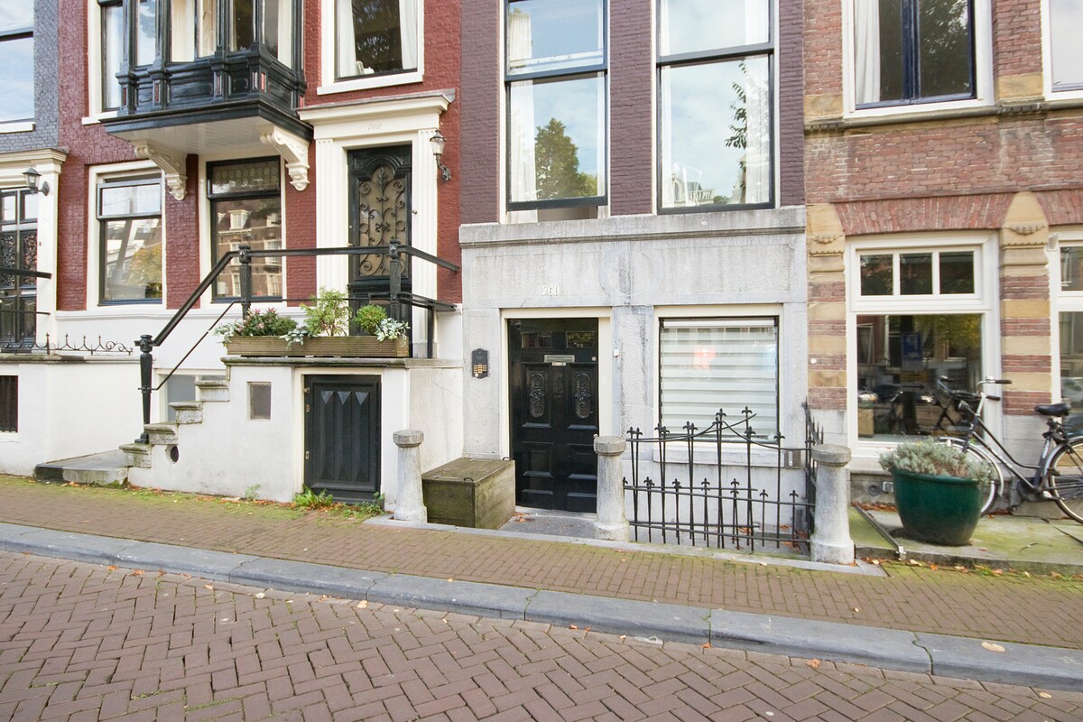 A charming canal house exterior is displayed, featuring brick walls and a dark wood entrance door adorned with ornate detailing. Steps lead up to the main entrance, flanked by greenery in planters, while a wrought iron gate encloses the property. Bicycles are parked nearby on the cobblestone street.