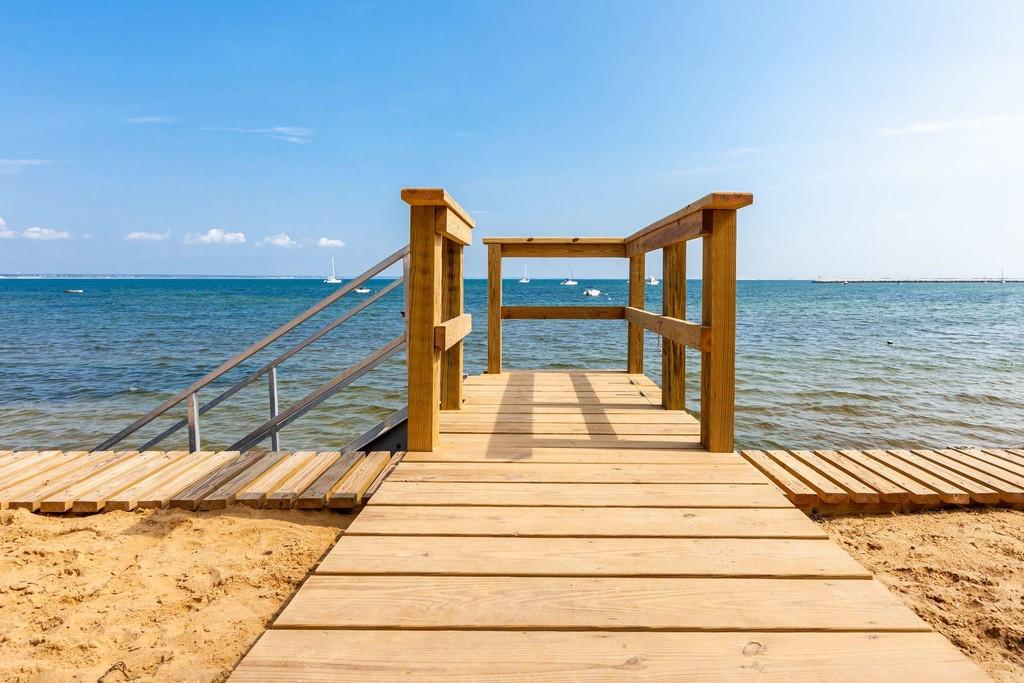A wooden walkway leads from the sandy beach to a dock extending over calm waters. The scene features a clear blue sky and distant sailboats on the bay, creating an inviting space for relaxation and enjoyment.