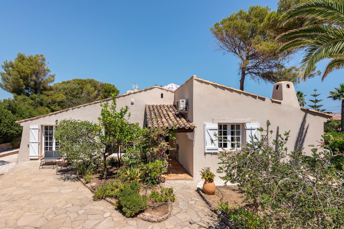 The villa features a light-colored exterior with a rustic tiled roof. Surrounding greenery and various plants enhance the inviting entrance, which includes white window shutters and a stone pathway leading to the door. A spacious outdoor area includes a harmonious blend of stone and flora.