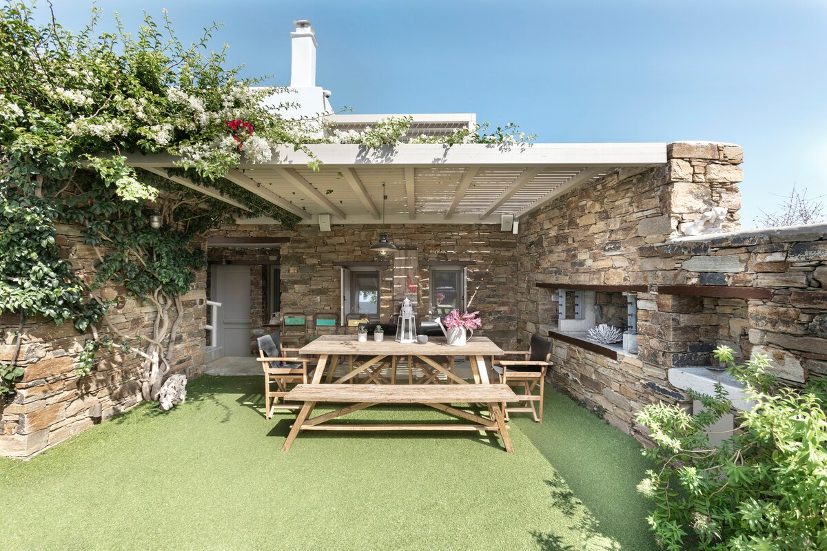 An outdoor dining area features a large wooden table surrounded by benches, all set on lush green grass. Natural stone walls and climbing greenery create a serene atmosphere, while light floods the space from above through an open roof.