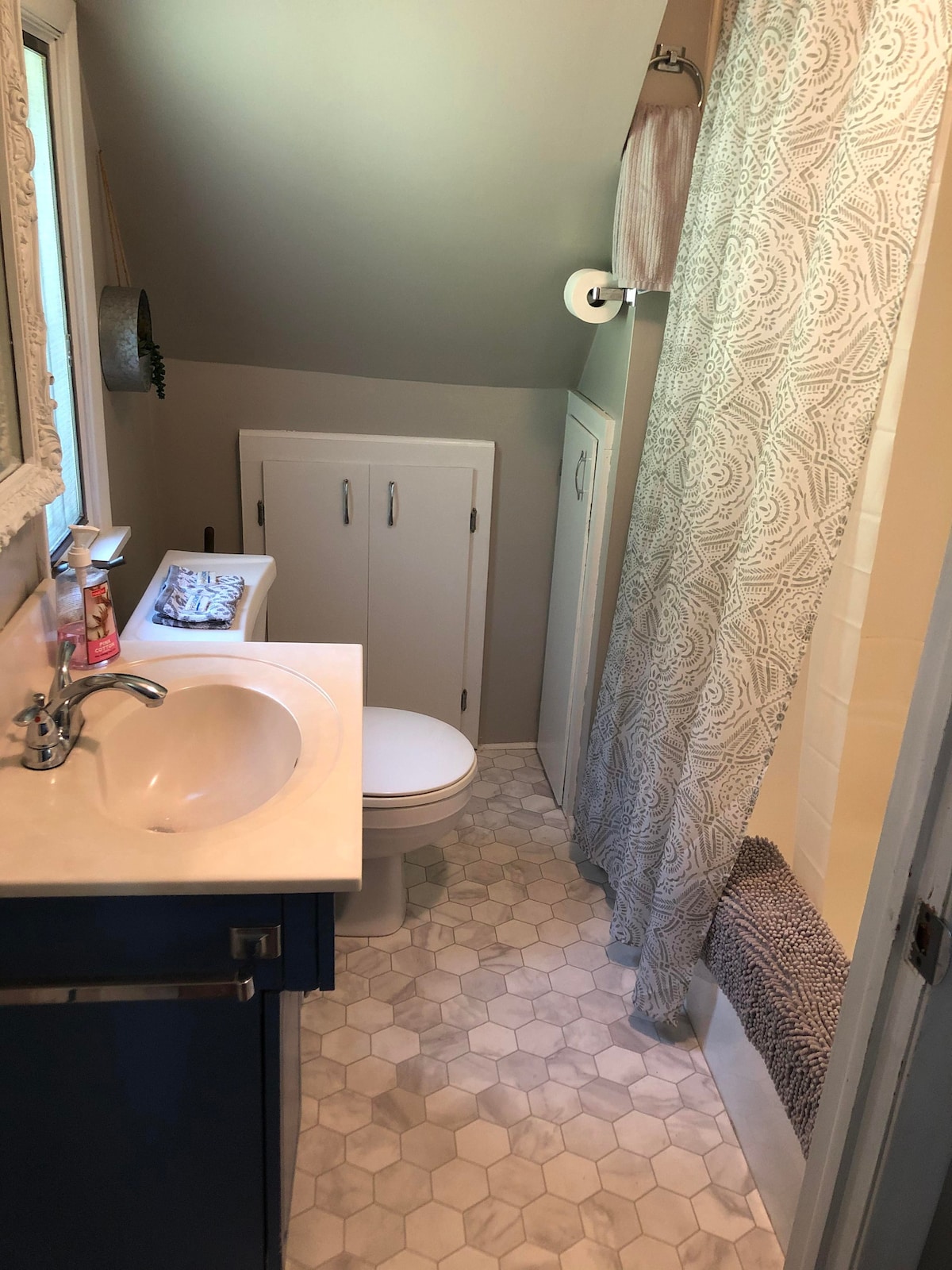 A well-appointed bathroom features hexagonal tile flooring and a light-colored vanity with a sink. A shower area is enclosed with a patterned curtain. Storage cabinets are positioned beside the sink, providing ample space for essentials. Natural light enters through a nearby window.