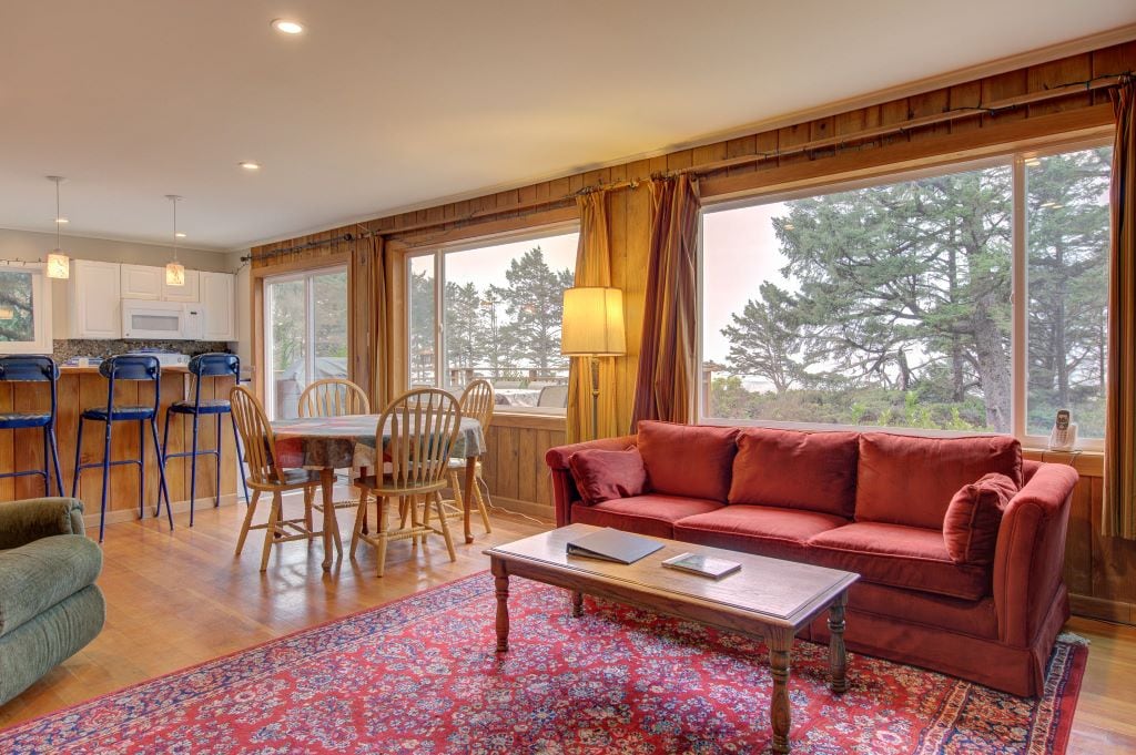 A warm living space showcases a deep red sofa and a wooden coffee table, complemented by a patterned rug. Large windows allow natural light and views of the surrounding greenery, with a dining area nearby featuring a wooden table and chairs, along with a breakfast bar in the background.