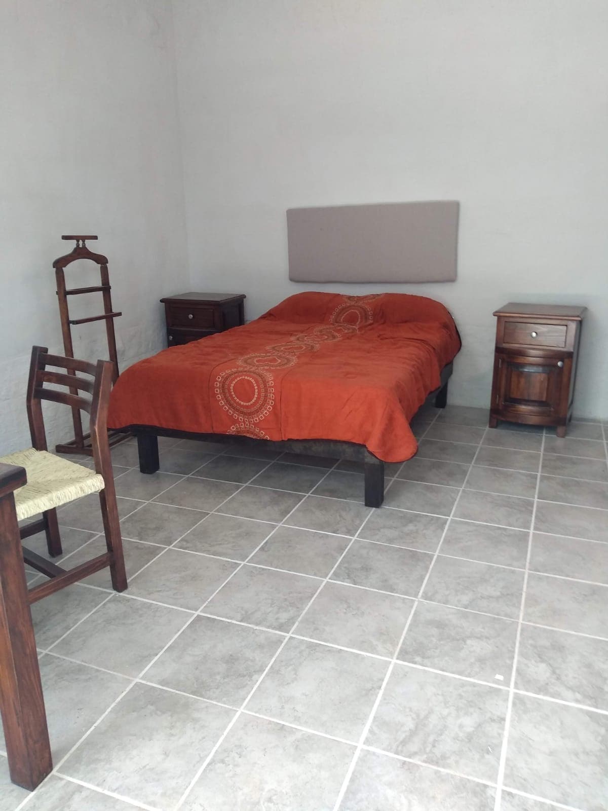 A cozy room is presented with a spacious bed covered in an orange bedspread. Dark wooden nightstands are positioned on each side of the bed, while a simple wooden chair and a small table are located to one side. The tiled floor adds a clean and open feel.