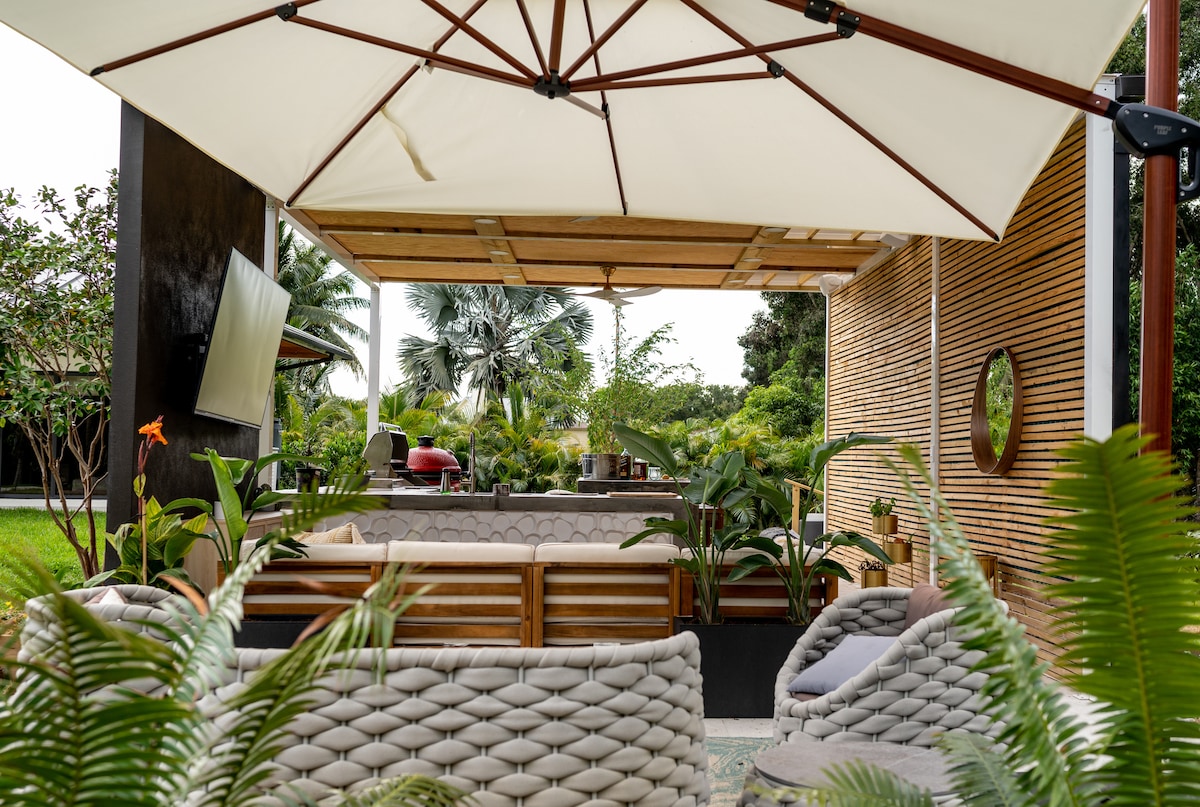A shaded outdoor seating area is presented beneath a large umbrella, featuring cozy wicker sofas surrounded by tropical greenery. A television is mounted on the wall, adding a modern touch to this inviting space, perfect for relaxation in the fresh air.
