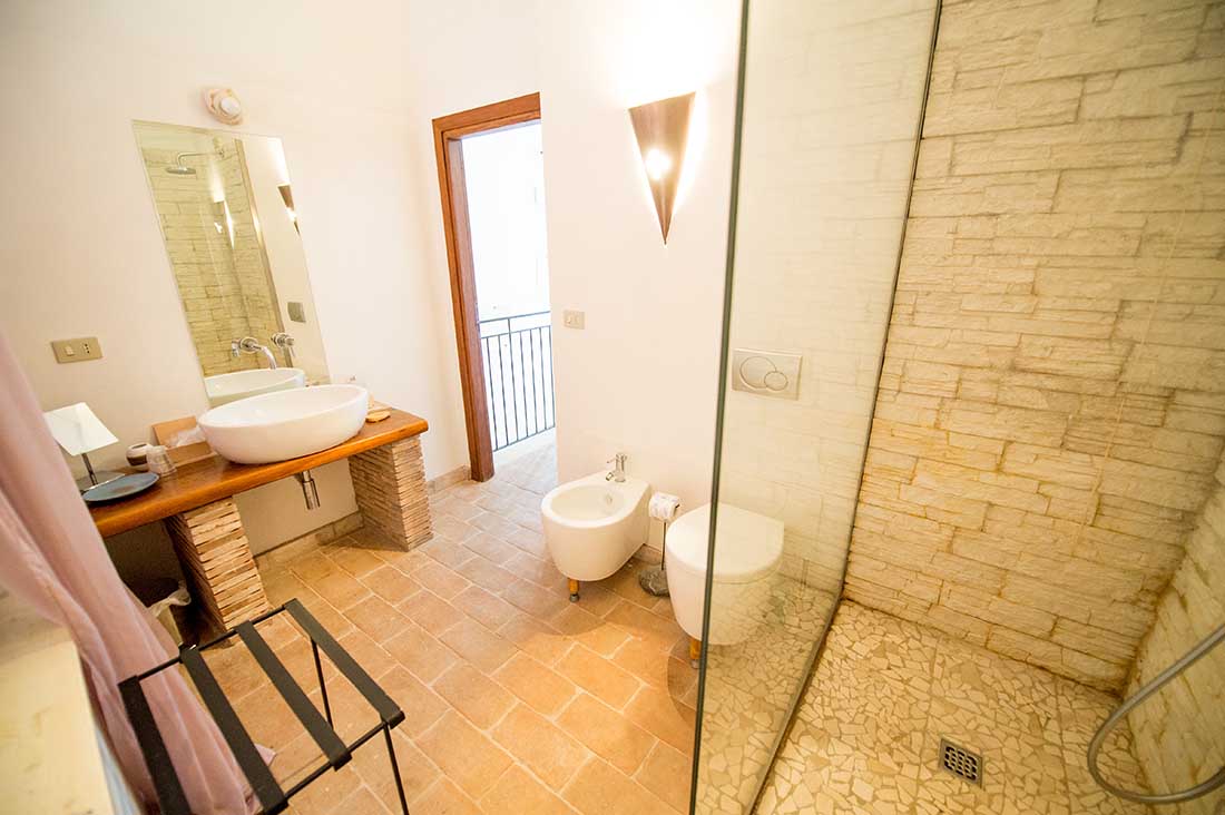 A bright bathroom is presented, featuring a walk-in shower with stone walls. A modern sink is mounted on a stone counter, accompanied by a bidet and toilet. Natural light enters through a window, and the floor is covered with terracotta tiles.