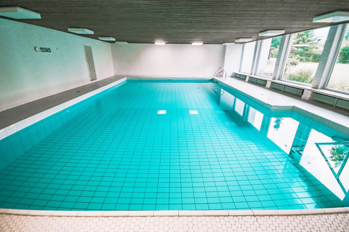An indoor swimming pool features turquoise tiles and is well-lit by overhead lights. Large windows allow natural light to illuminate the space, providing views of the surrounding landscape. A ladder leads into the water, enhancing accessibility.