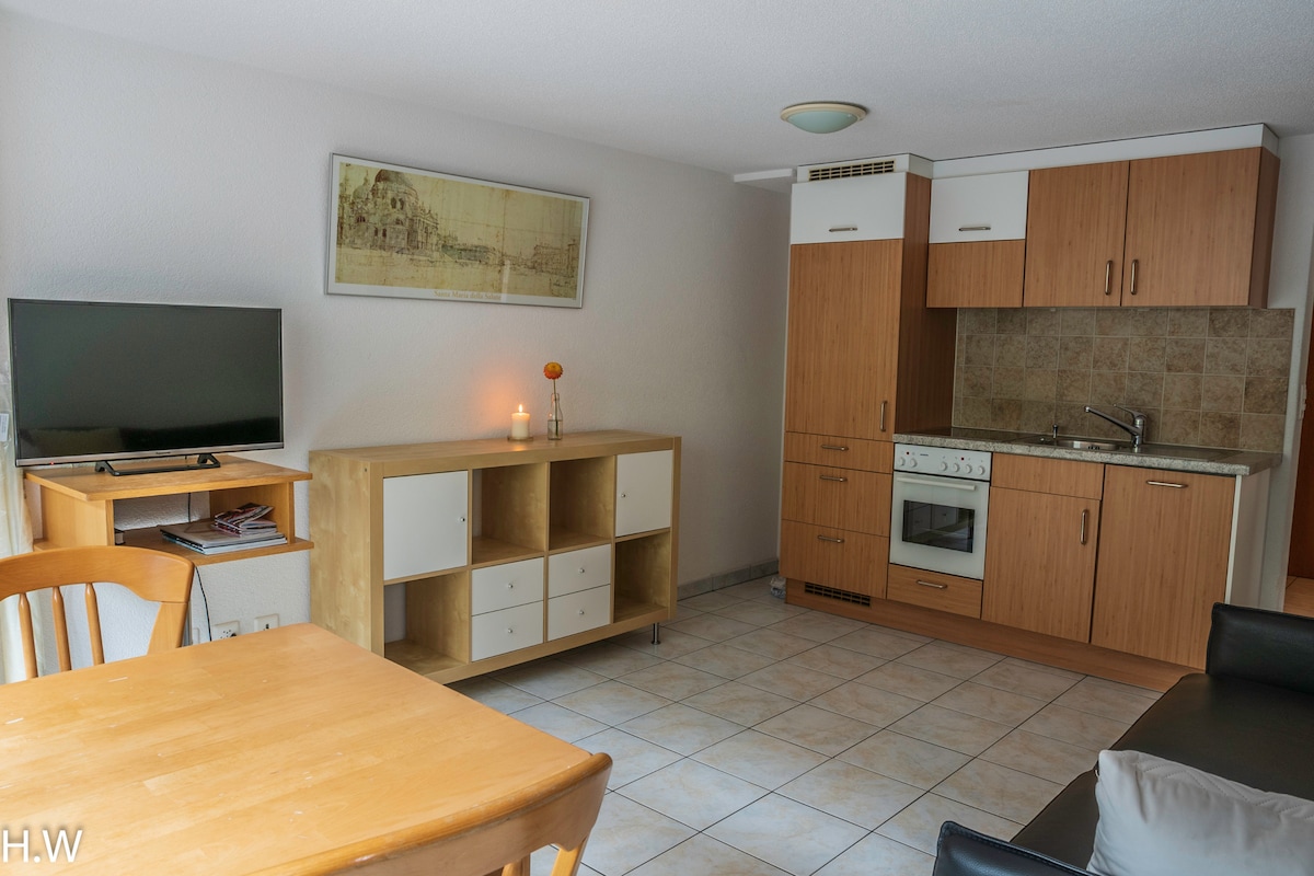 A light-filled living area is shown, featuring a dining table with wooden chairs and a compact kitchen area with built-in cabinets. A TV stands on a wooden shelf, and soft lighting enhances the neutral color scheme of the walls and tiled flooring.
