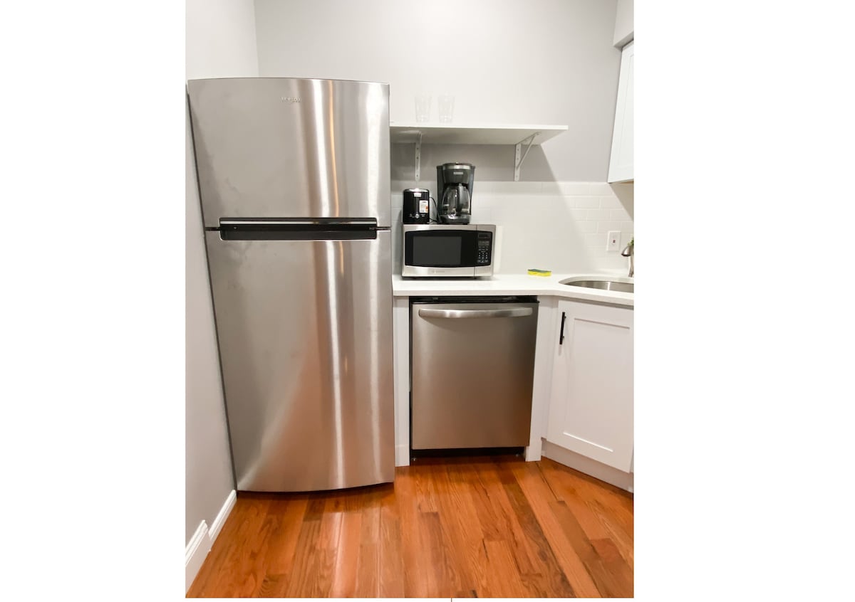 A modern kitchen area features stainless steel appliances, including a refrigerator, microwave, and dishwasher. A coffee maker is positioned on the counter, complemented by white cabinetry and a clean countertop. The space is illuminated by overhead lighting that enhances the warm wooden flooring.