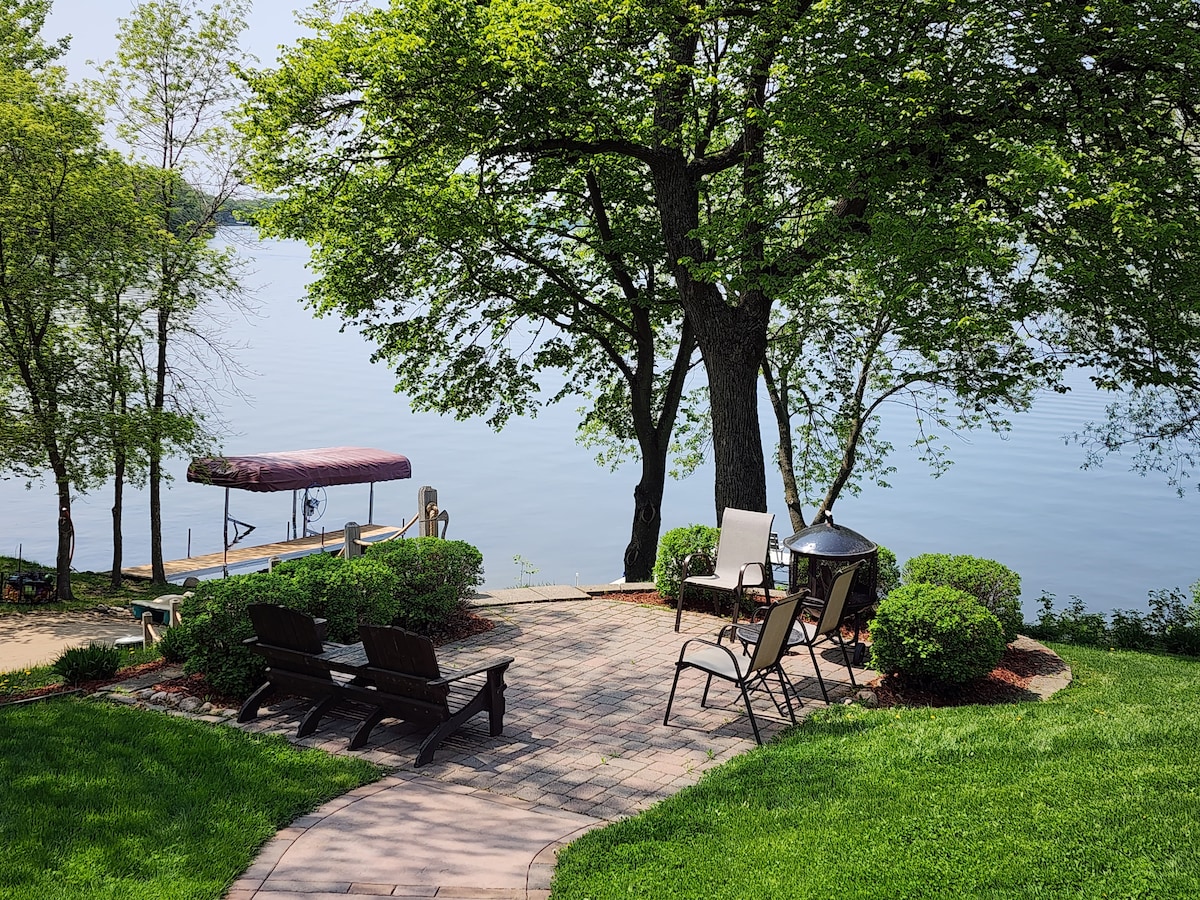A tranquil outdoor space features a circular paved patio surrounded by manicured grass and shrubs. Comfortable seating areas are arranged among the greenery, with a lakeside view of Diamond Lake visible in the background and a covered dock nearby.