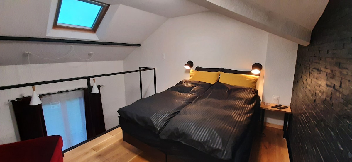 A spacious mezzanine sleeping area features a large bed with dark bedding. Two bedside lamps provide soft lighting, while a window with a skylight allows natural light to brighten the room. A textured dark wall serves as an elegant backdrop.