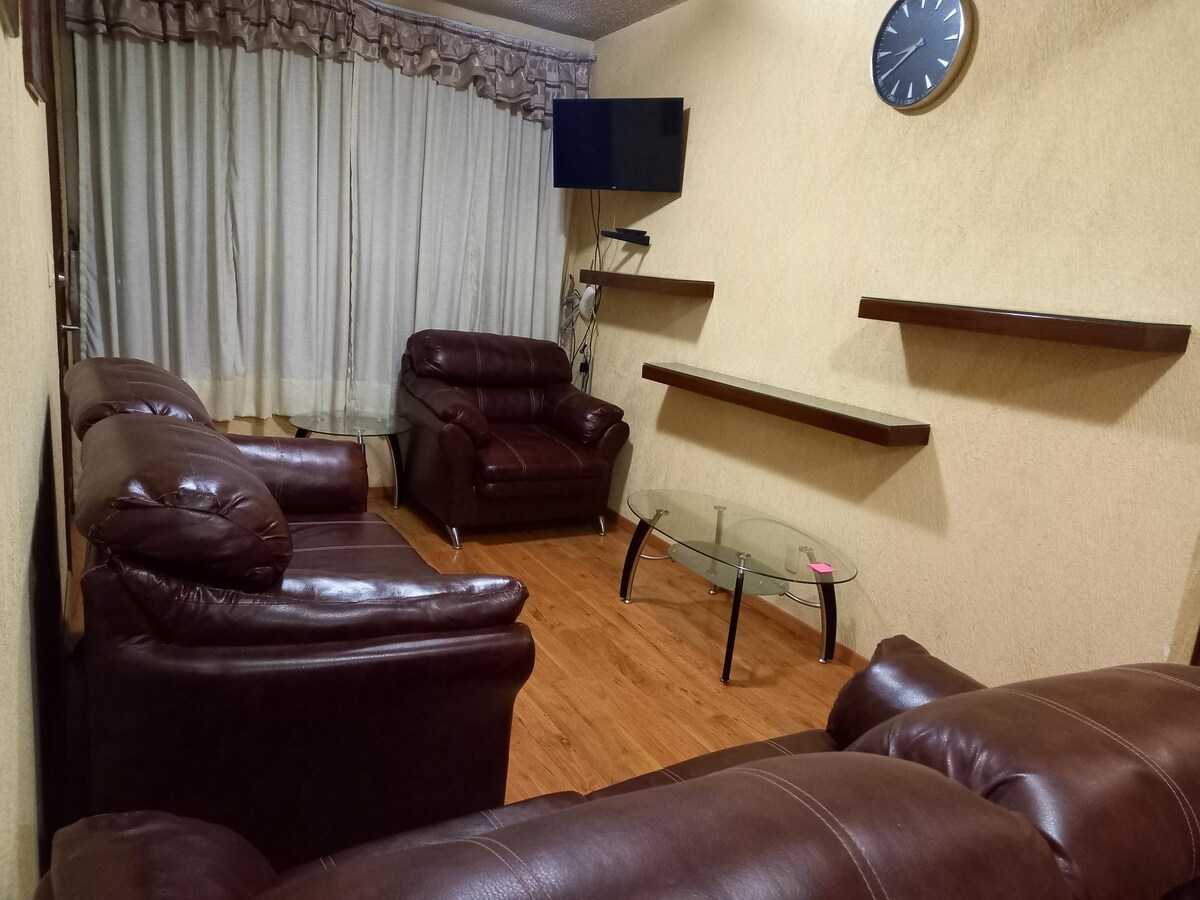 A living area features a combination of comfortable brown leather seating, including two armchairs and a sectional couch. A glass coffee table is positioned in the center, with a wall-mounted television and shelves providing functional decor. Natural light filters through sheer curtains.