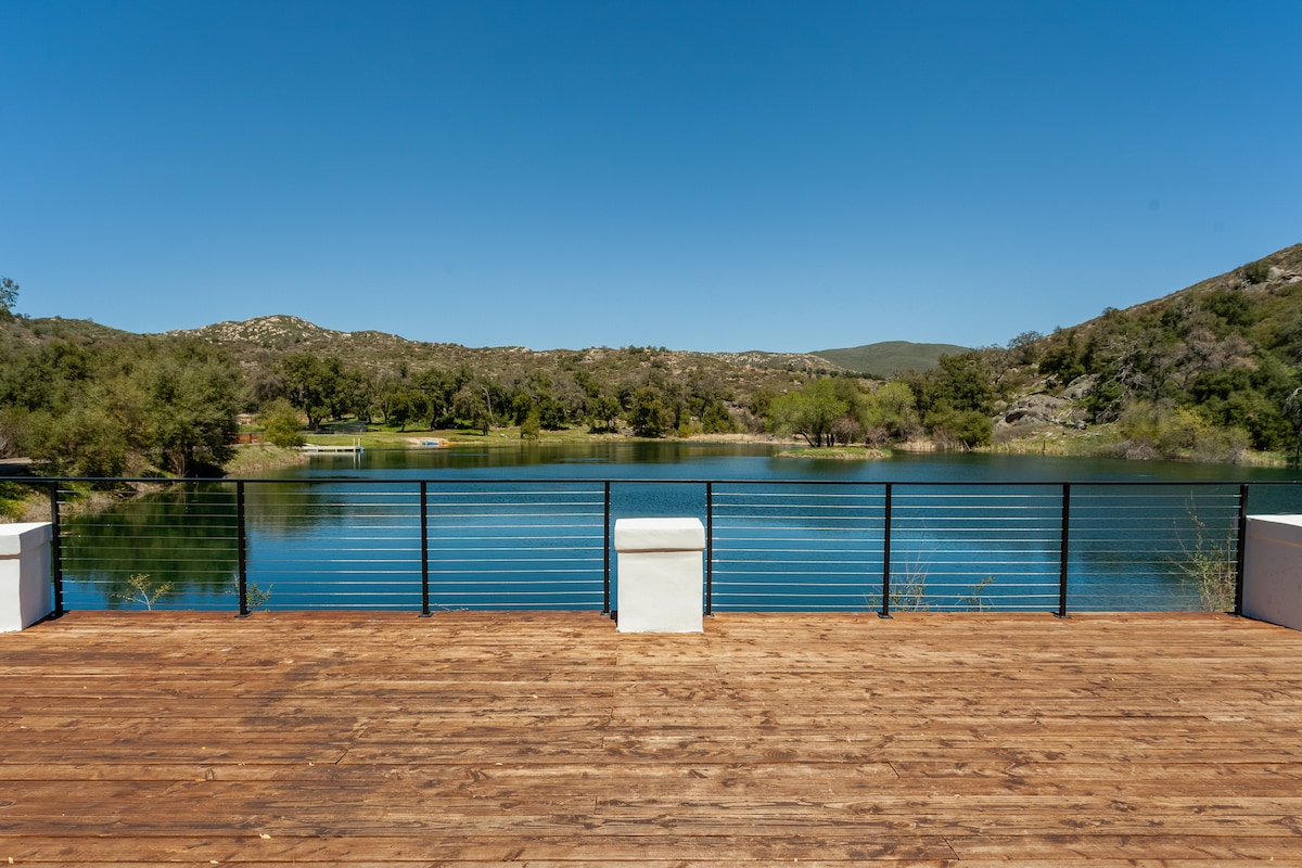 A spacious wooden deck extends over the water, providing a tranquil view of the lake and surrounding greenery. The railing offers safety while allowing for unobstructed sightlines of the calm water and distant hills. Clear skies create a serene atmosphere.