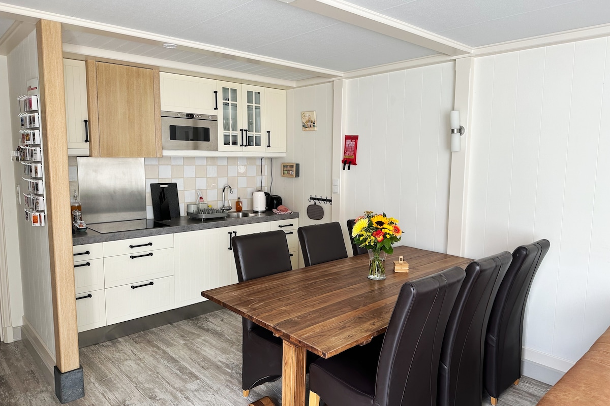 A modern kitchen area is shown, featuring light-colored cabinets and a stainless steel refrigerator. A large wooden dining table surrounded by six black leather chairs is visible, along with a bouquet of sunflowers as a centerpiece. Cooking appliances are neatly arranged on the countertop.