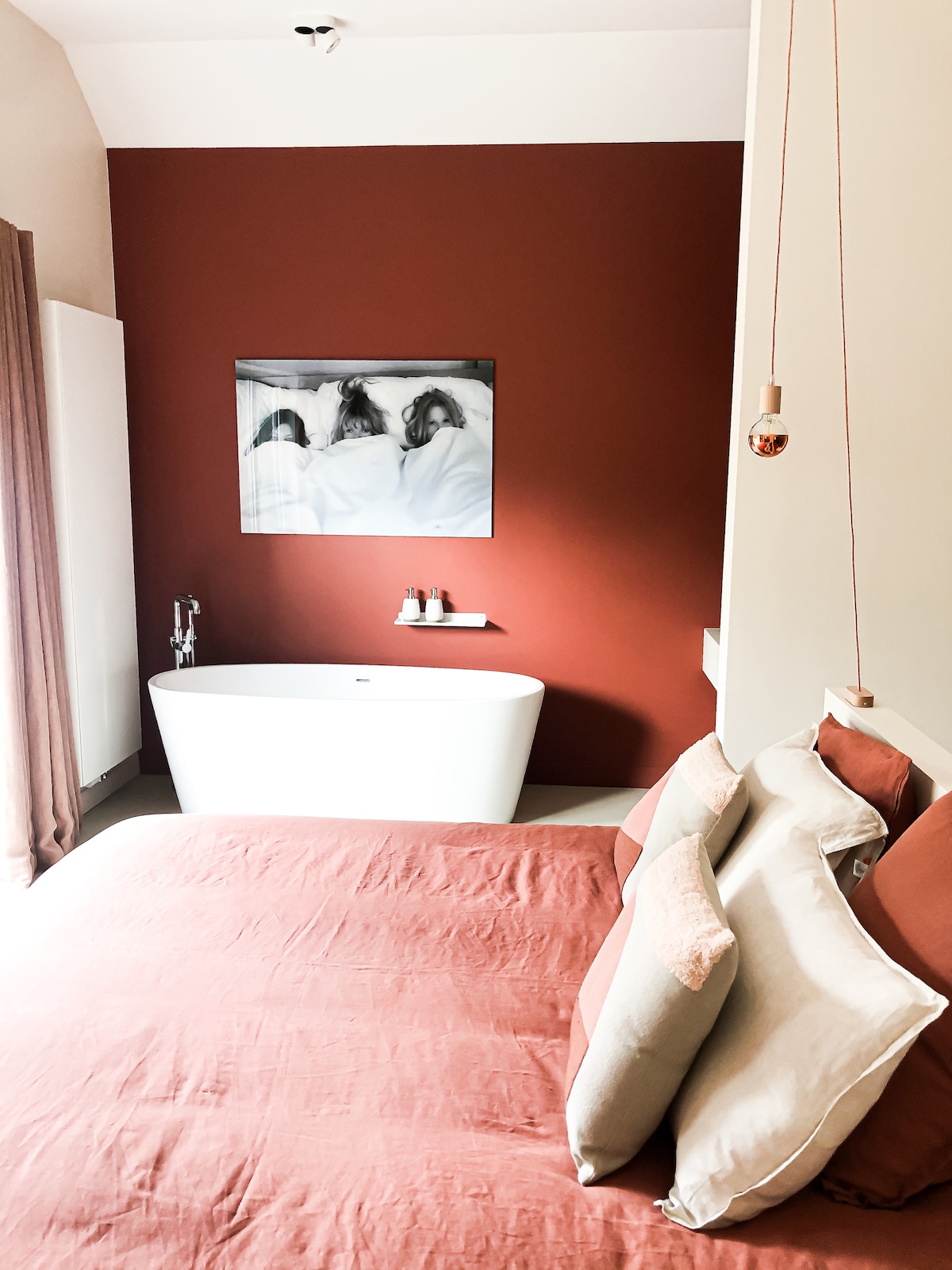 A cozy bedroom is highlighted by a warm red accent wall. A spacious double bed with various decorative pillows sits at the forefront. A modern freestanding bathtub is situated adjacent, with a framed artwork mounted above. Soft curtains drape by a window, adding to the room's tranquility.