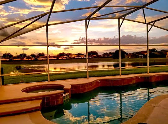 The screened lanai pool area is highlighted by a spacious pool and spa, reflecting the vibrant sunset. A serene lake and green yard are visible beyond the enclosure, providing a peaceful backdrop.