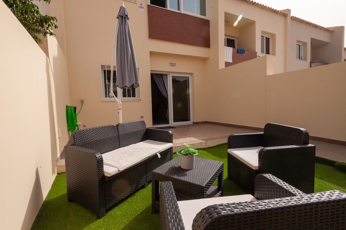 A private outdoor terrace features comfortable seating arranged around a low table, set on a grassy area. A patio umbrella provides shade, while the surrounding walls ensure privacy. In the background, large glass doors lead into the interior space.