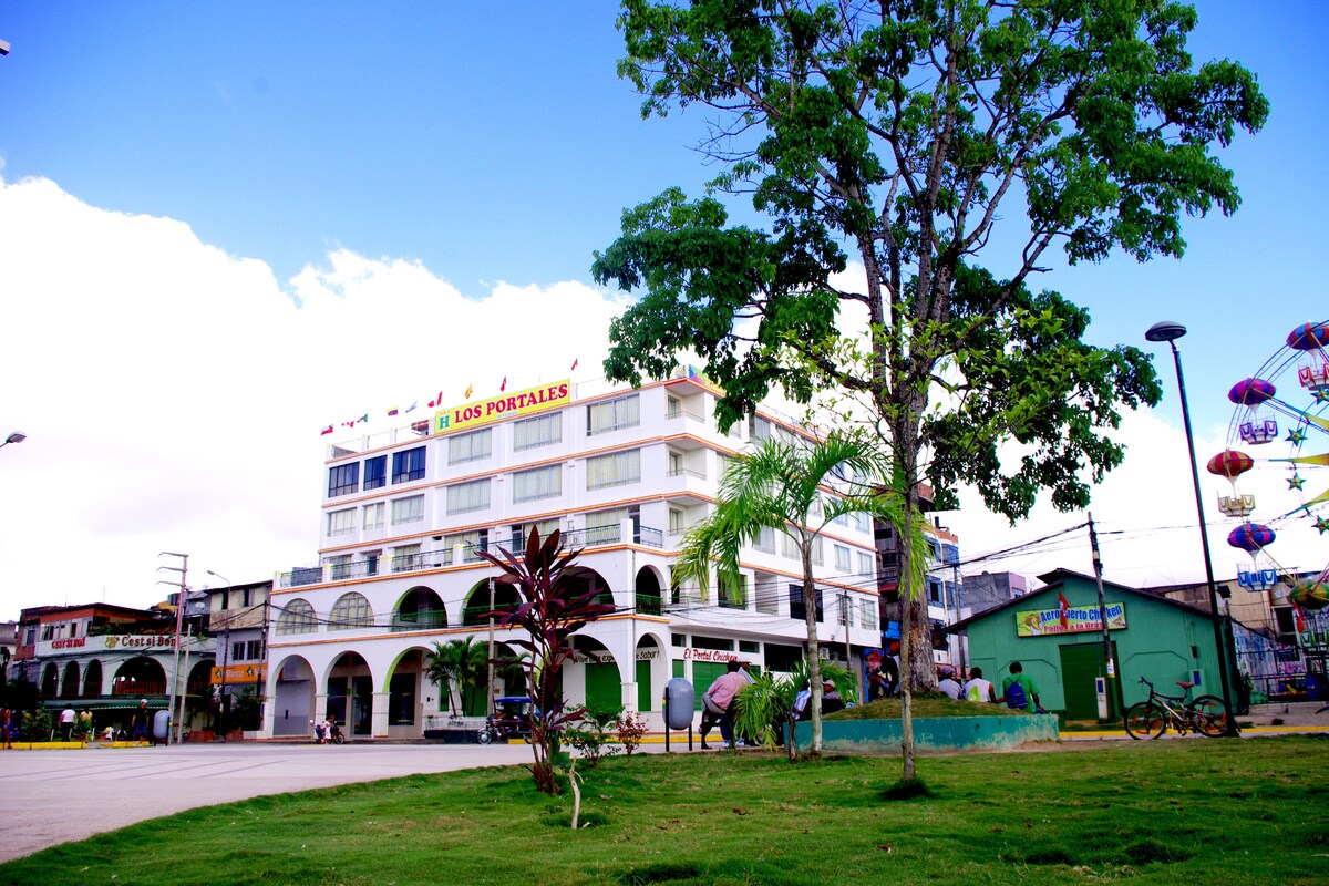 The hotel is showcased in a vibrant urban setting, surrounded by green grass and decorative trees. The multi-story building features large windows and balconies, with colorful signage that identifies the establishment. Nearby, local shops and entertainment options are visible, enhancing the area's lively atmosphere.