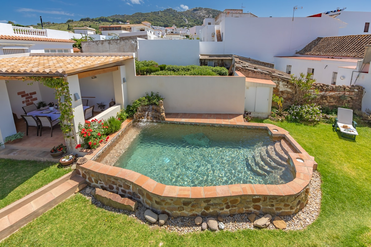 A serene swimming pool is surrounded by lush green grass and flowering plants. The pool features a natural stone edge and a gradual step entry. Nearby, a shaded seating area offers a table and chairs for relaxation, complemented by a view of surrounding white-walled buildings.