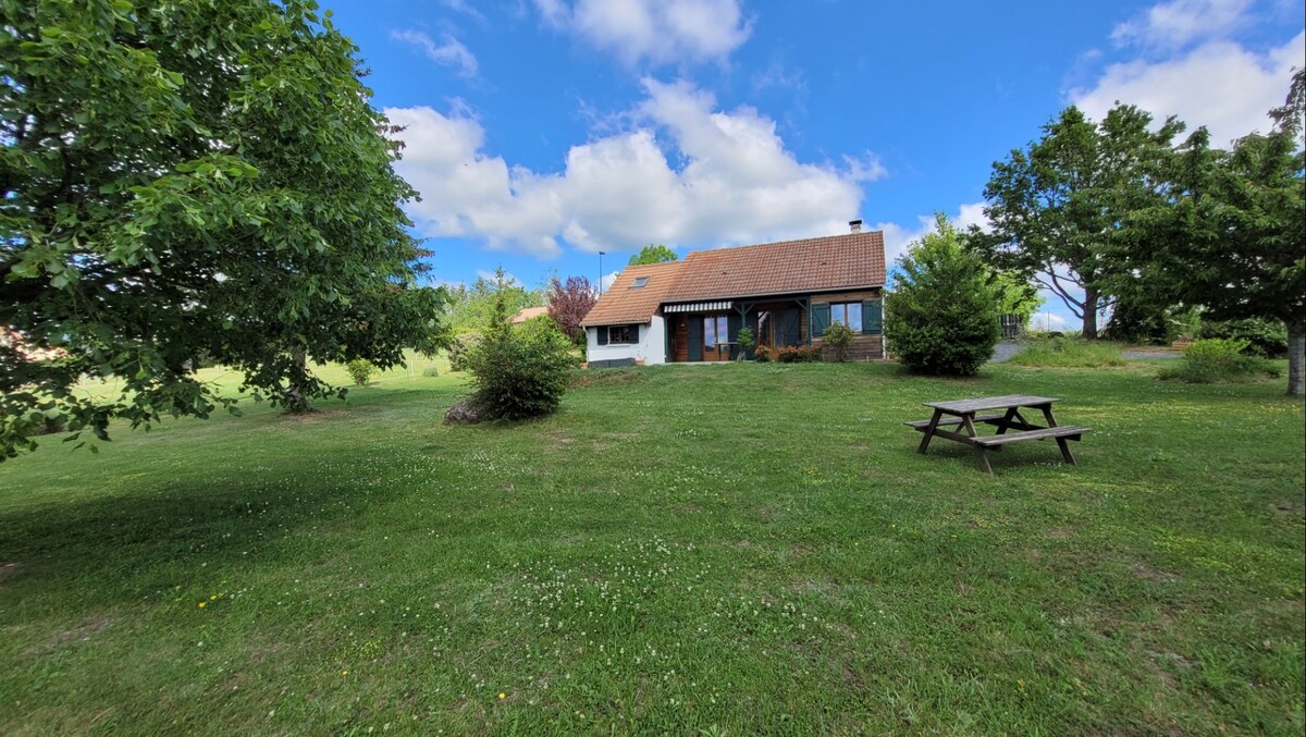 A single-story house is set on a spacious lawn, surrounded by trees and greenery. A picnic table is positioned near the house, providing an inviting outdoor space. The sky is partially cloudy, with soft blue tones enhancing the tranquil atmosphere.