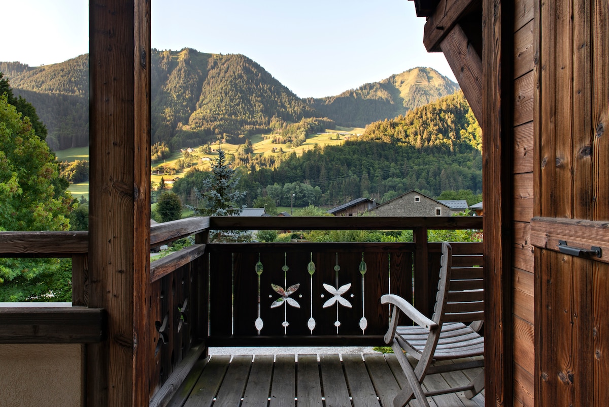 A wooden balcony offers a view of rolling green hills and mountains in the distance. The structure features rustic wood railings with decorative cutouts, while a simple chair invites relaxation in the serene alpine surroundings.