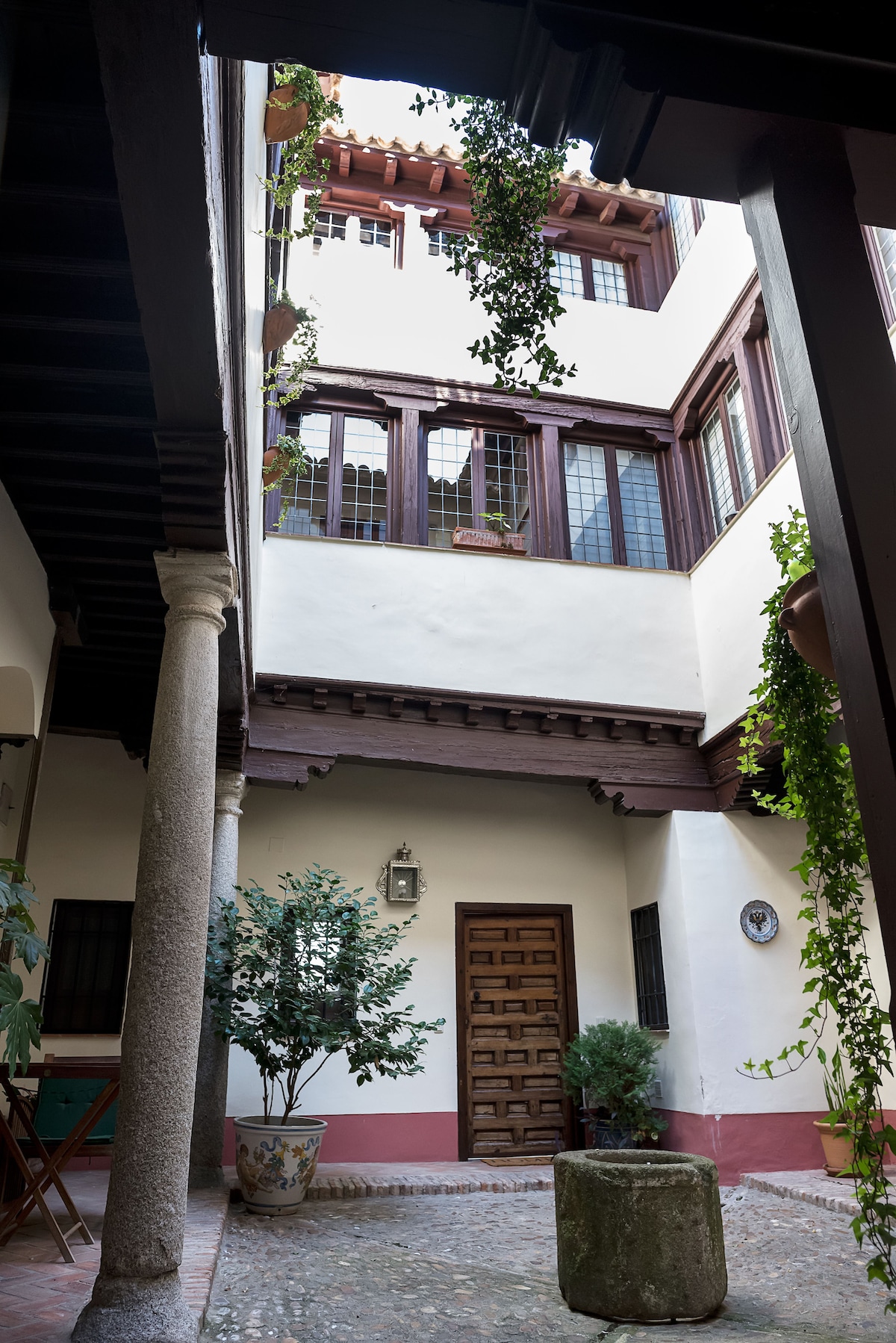 A historic courtyard features traditional wooden beams and terracotta tiles, with lush greenery cascading from balconies. A round planter sits in the center, surrounded by a stone pathway. The walls display a mix of white and muted tones, enhancing the charm of the architecture.