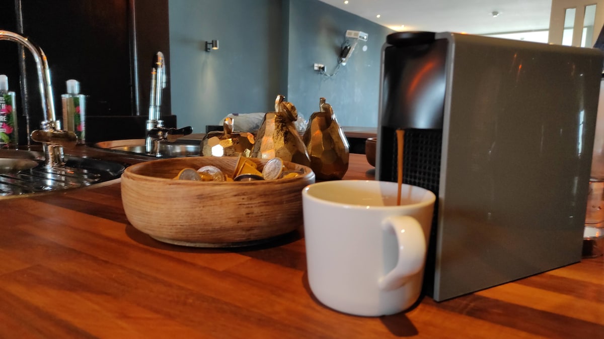 A modern coffee machine sits on a polished wooden countertop, pouring coffee into a white mug. A bowl containing various coffee capsules and decorative golden figures are positioned nearby, enhancing the functional yet stylish kitchen environment.