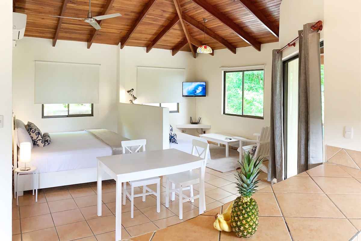 A spacious studio is presented with a high wood-beam ceiling. Natural light illuminates the area, featuring a spacious bed and a dining table with white chairs. A television is mounted on the wall, with a cozy seating arrangement nearby. A kitchenette and fresh fruit are placed on a tiled counter.