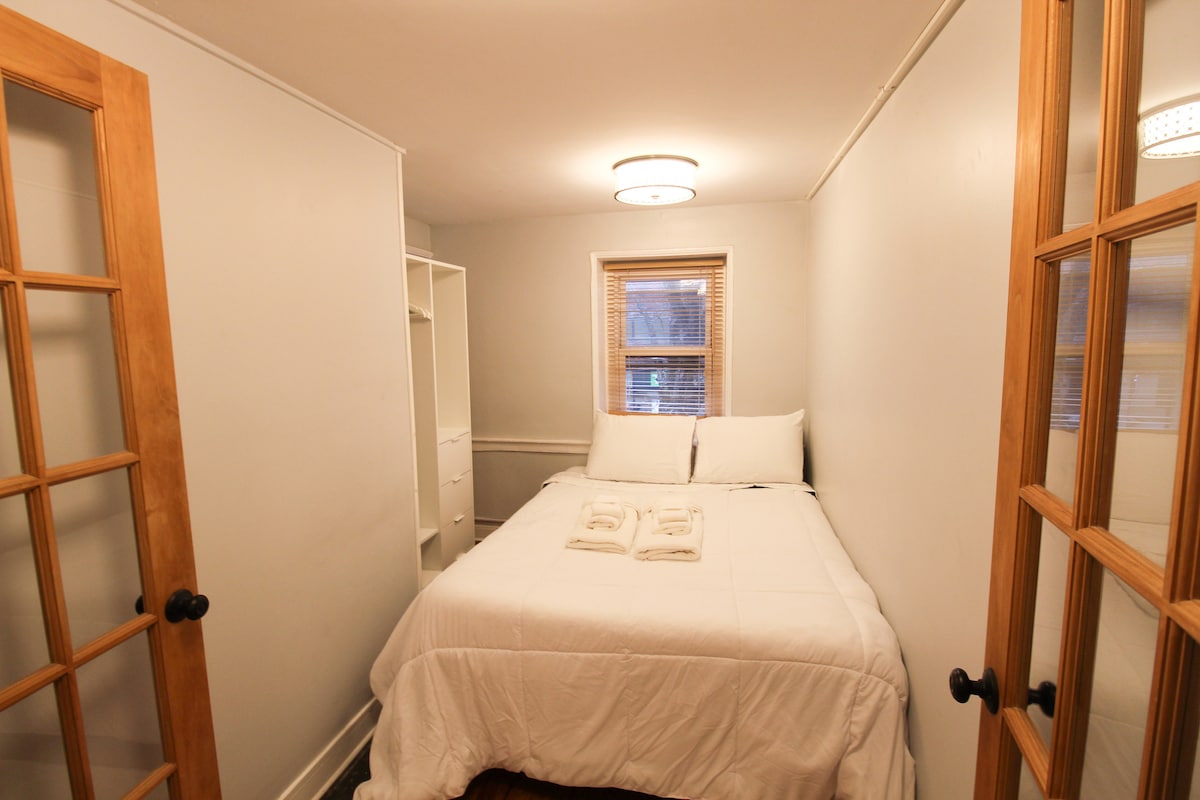 A small room is observed, featuring a spacious bed dressed in white linens with two folded towels placed atop. Natural light enters through a window, while built-in shelving provides storage options. French doors offer access to the room, enhancing the overall minimalist design.