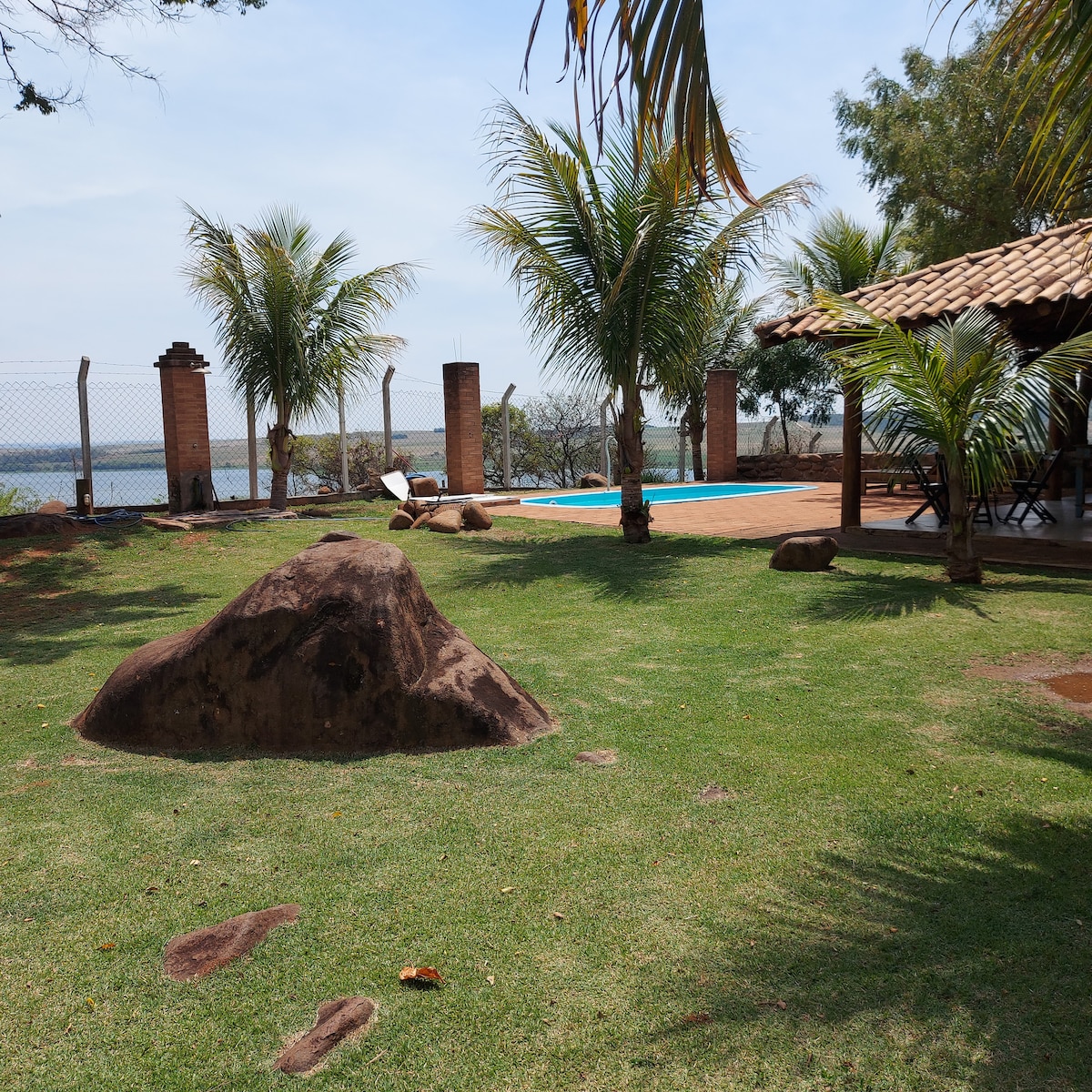 The outdoor area features a well-maintained lawn dotted with natural stones and palm trees. A wooden deck houses a poolside lounge area, with a swimming pool visible in the distance against the backdrop of the reservoir, creating a serene escape.