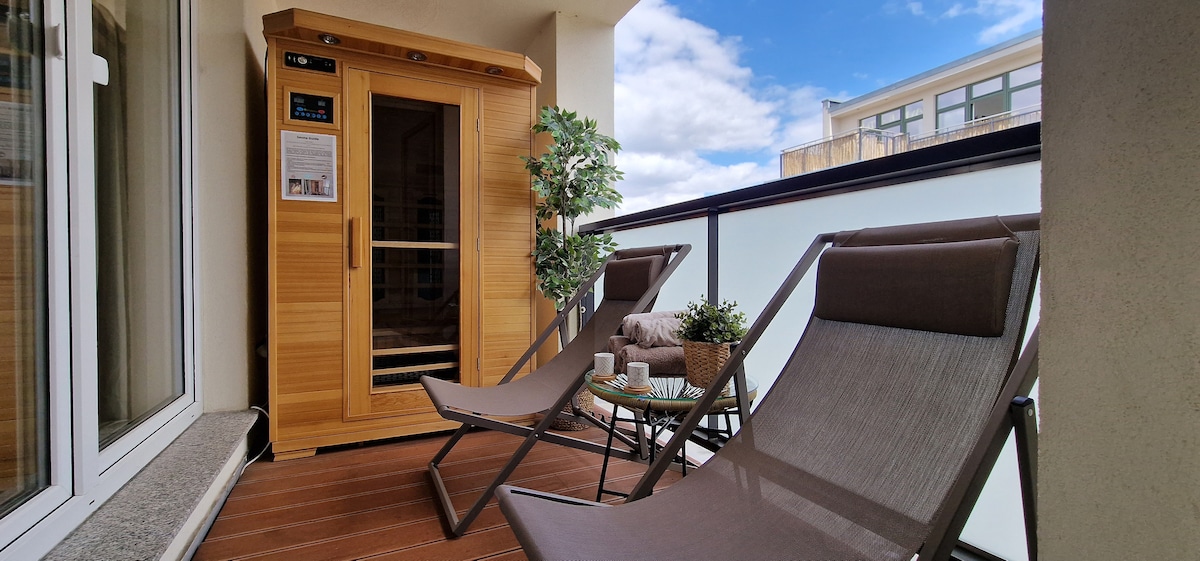 A private balcony features a wooden infrared sauna, accompanied by two relaxing lounge chairs. A small table is set with tea and coffee supplies, while a decorative plant adds a touch of greenery. Light clouds drift across a blue sky overhead.