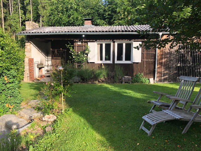 A charming exterior view of the tiny house is set against a backdrop of lush greenery. The structure features a mix of wooden and paneling, with several windows allowing natural light. A well-maintained garden surrounds the house, and wooden lounge chairs are positioned on the lawn.