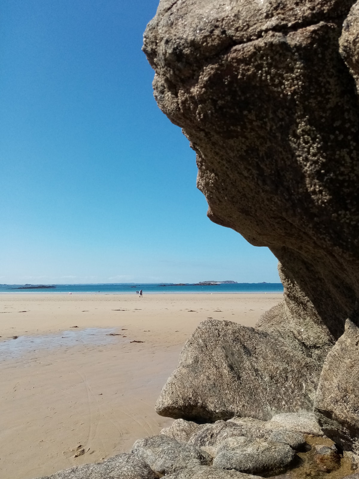 A view of the beach shows a sandy shoreline stretching towards the horizon, where the ocean meets a clear, blue sky. In the distance, a couple of people can be seen walking along the water's edge, framed by a large rock formation in the foreground.