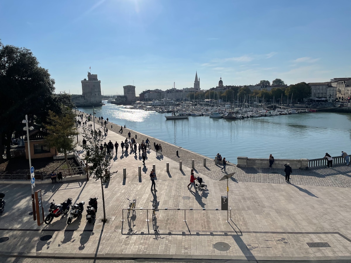 A lively waterfront scene along the Vieux Port is visible, showcasing a promenade filled with pedestrians. Boats are anchored in the calm water, with historic buildings and greenery framing the view. The clear sky and sunlight enhance the vibrant atmosphere.