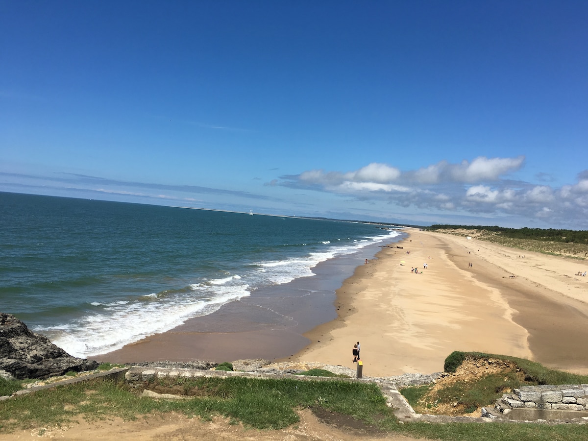The image captures a wide sandy beach bordered by gentle waves and a clear blue sky. The shoreline extends into the distance, with patches of grass and rocks visible in the foreground. A few distant figures can be seen along the beach.