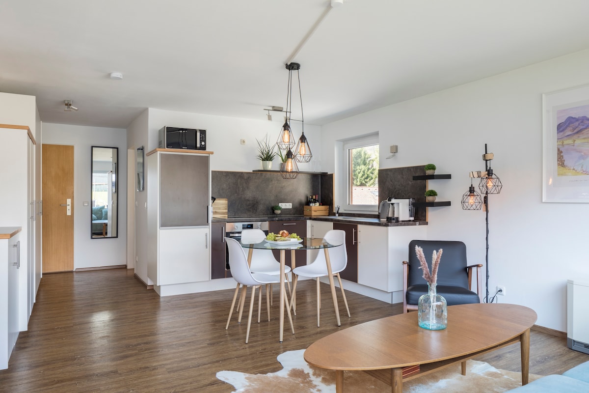 An open living area showcases a modern kitchen with sleek cabinetry, a dining table set for four, and a comfortable seating arrangement. Warm wood flooring complements the neutral tones, while natural light flows through a window, highlighting decorative plants and stylish lighting fixtures.