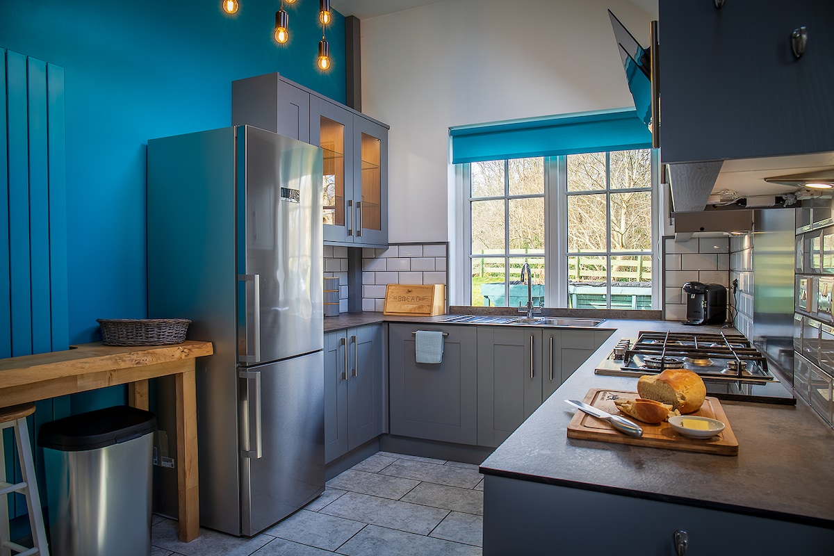 A modern kitchen features sleek cabinetry in gray tones and a blue accent wall. Stainless steel appliances are present, including a refrigerator and a gas stove. Natural light filters through large windows, illuminating a countertop with cooking utensils and baked goods.