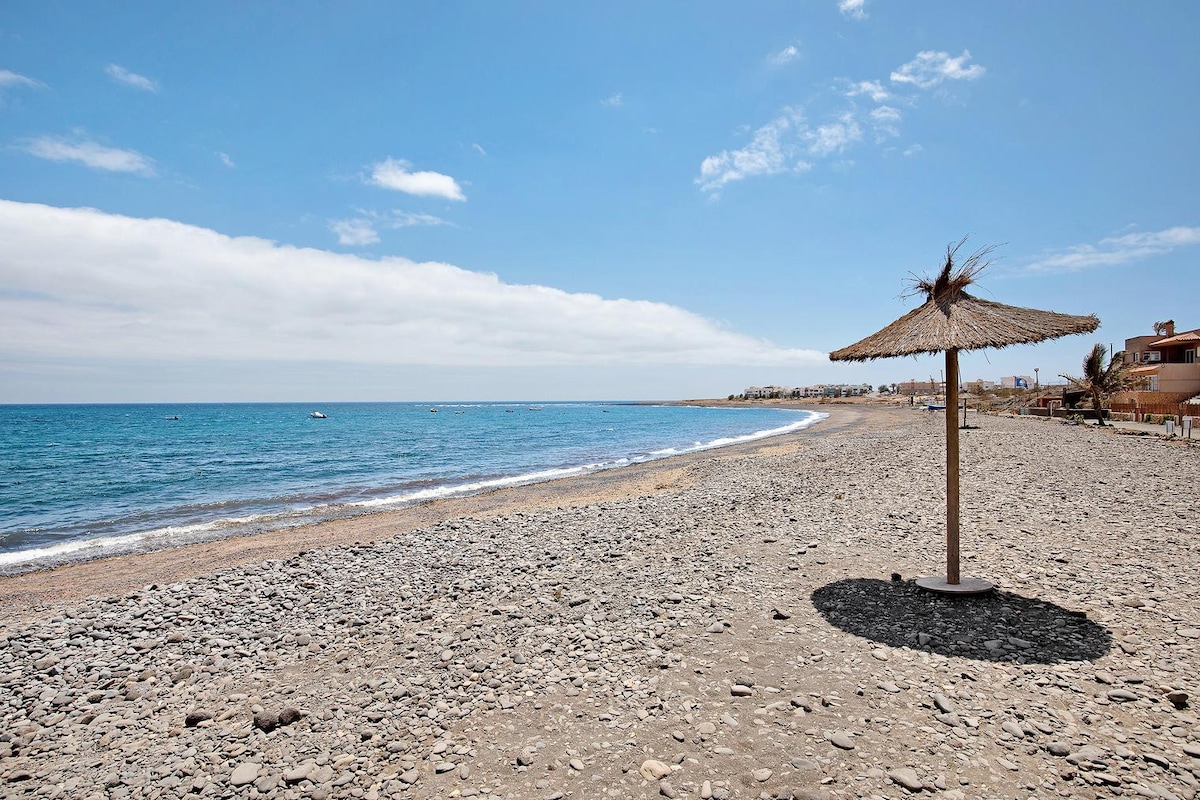 A scenic beach scene is captured, featuring a rocky shoreline that meets the calm blue waters. A straw umbrella provides shade, casting a shadow on the pebbled beach. The clarity of the sky and water enhances the sense of tranquility.