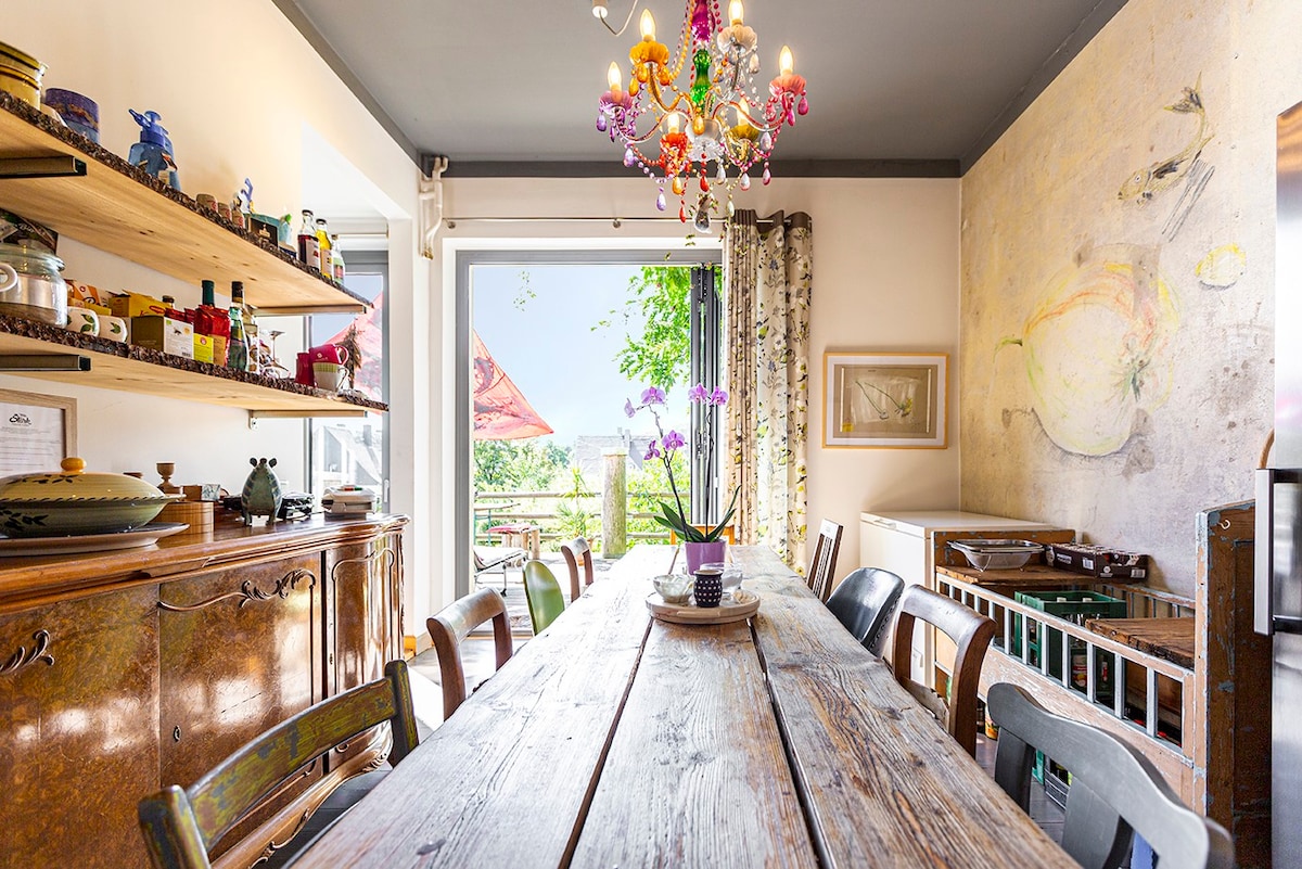 A spacious dining area is highlighted by a long wooden table surrounded by multiple chairs. A chandelier adds a touch of elegance, while large windows provide natural light and a view of the outdoors. Shelves display various decorative items and kitchen essentials.