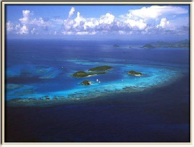 An aerial view captures a cluster of small islands surrounded by vibrant blue water. The landscape features varying shades of turquoise and deep blue, with coral reefs visible beneath the water's surface, and scattered clouds dot the sky.