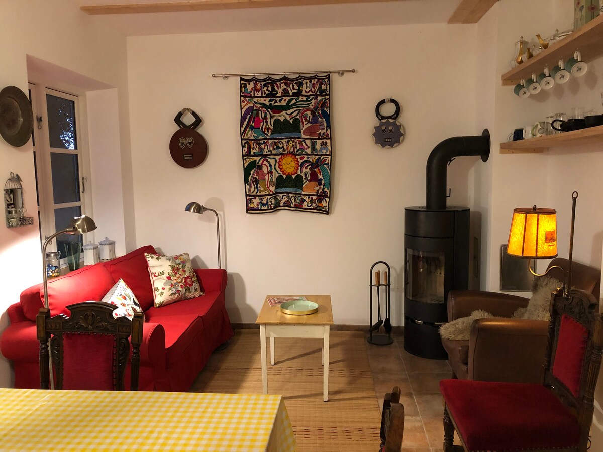 A cozy living area is highlighted by a bright red couch adorned with decorative pillows. A rustic wooden coffee table sits in front of a black stove, with light emanating from a nearby lamp. A colorful tapestry hangs on the wall, contributing to the room's warmth.