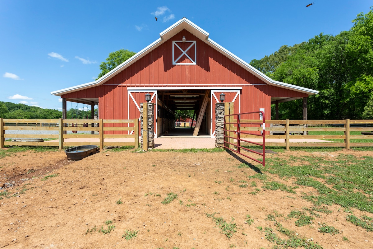 A spacious red barn with white trim and a large entrance is set against a clear blue sky. The barn's open structure allows for direct access to the surrounding yard and fields, framed by a wooden fence and vibrant greenery.