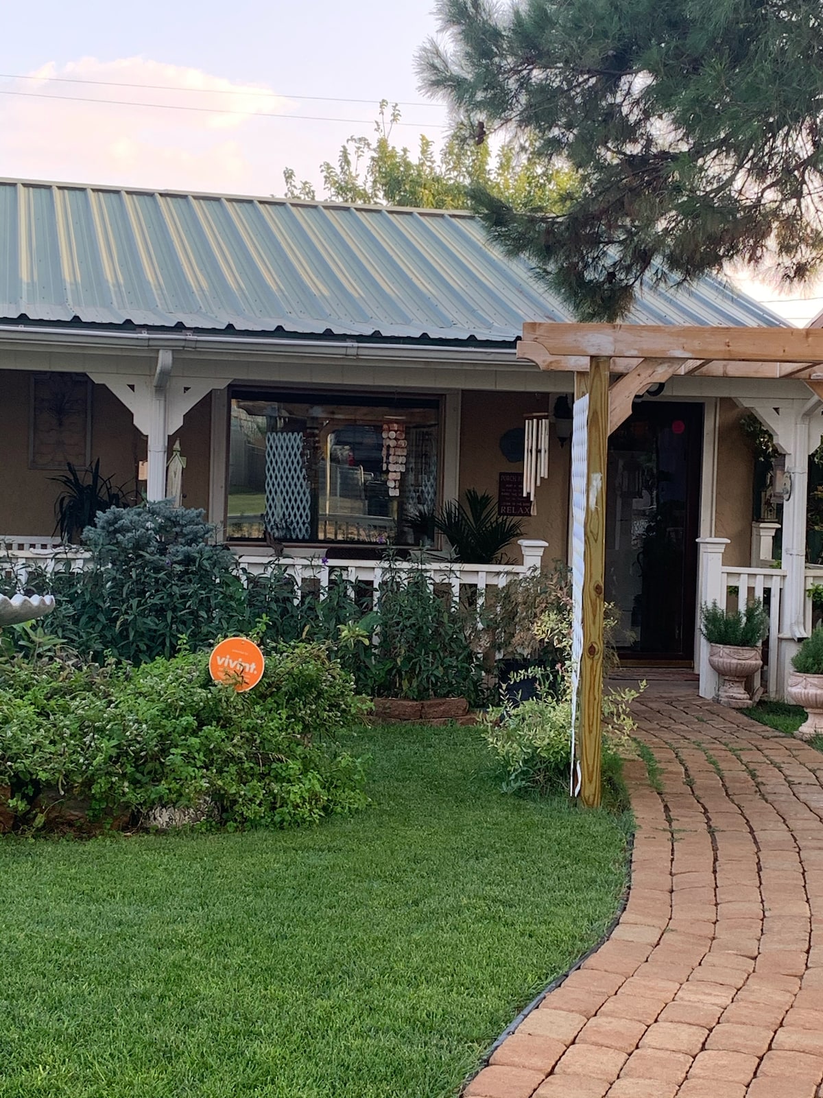 A charming entrance is framed by a lush front garden featuring green shrubs and flowering plants. A stone pathway leads to the welcoming porch, which is covered by a wooden trellis. The exterior displays a metal roof and warm colors, enhancing its inviting appeal.