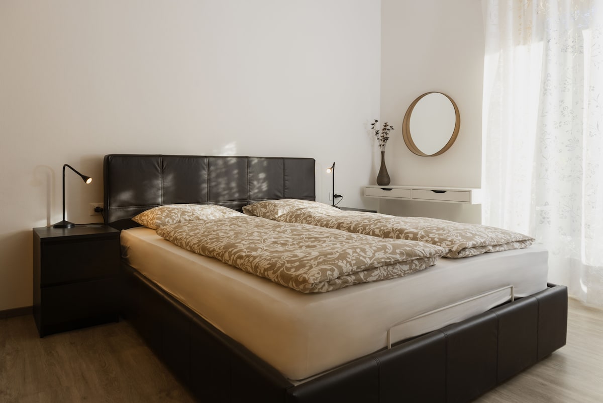 A spacious bed with a neutral-colored bedding set occupies the center of the room. Two bedside lamps are placed on dark nightstands. Natural light filters through sheer curtains, casting soft shadows, while a round mirror hangs on the wall, enhancing the room's brightness.