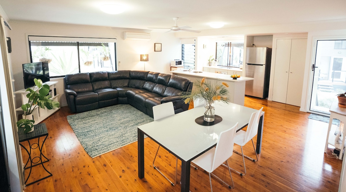 The open living area features a large, dark leather sectional sofa, complemented by a glass dining table with white chairs. Natural light illuminates the space through the large windows, highlighting the timber floors and minimalistic decor while a ceiling fan circulates air above.