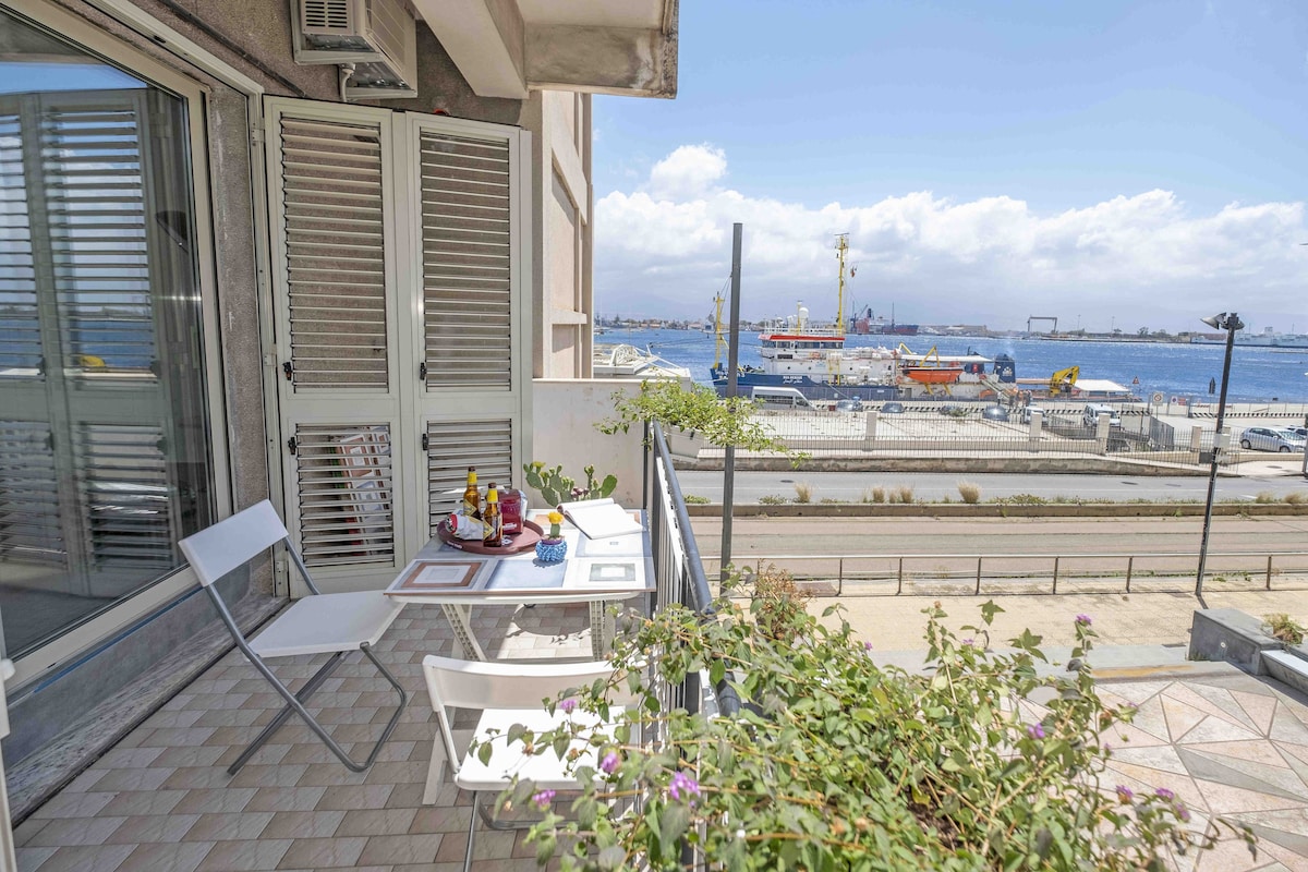 A small balcony features two white chairs and a table set for breakfast, overlooking the harbor. Vibrant plants line the railing, and large ships can be seen in the distance, with clear skies and gentle clouds providing a scenic backdrop.