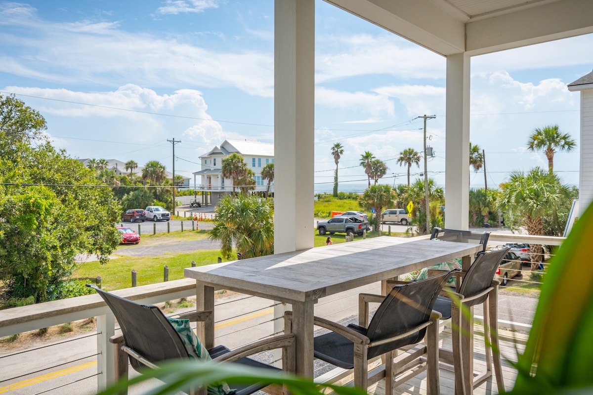 An outdoor seating area features a long wooden table surrounded by four cushioned chairs, positioned on a covered porch. The view includes palm trees and glimpses of the beach in the distance, with vehicles parked along the road in the foreground.