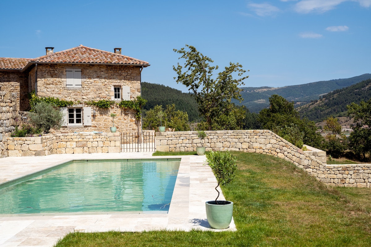 The image captures a renovated stone building nestled on a hillside, surrounded by verdant landscapes. A sparkling swimming pool is visible in the foreground, featuring a tiled deck and bordered by neatly trimmed grass. The backdrop showcases a panoramic view of rolling hills beneath a clear blue sky.