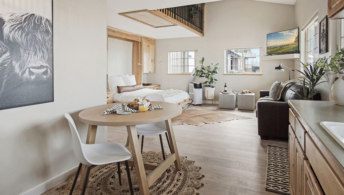 An open living area is presented, featuring a round dining table with two chairs and a cozy sofa. A comfortable queen bed is positioned in the background. Natural light filters through two windows, illuminating the warm wooden accents and soft neutral tones throughout the space.