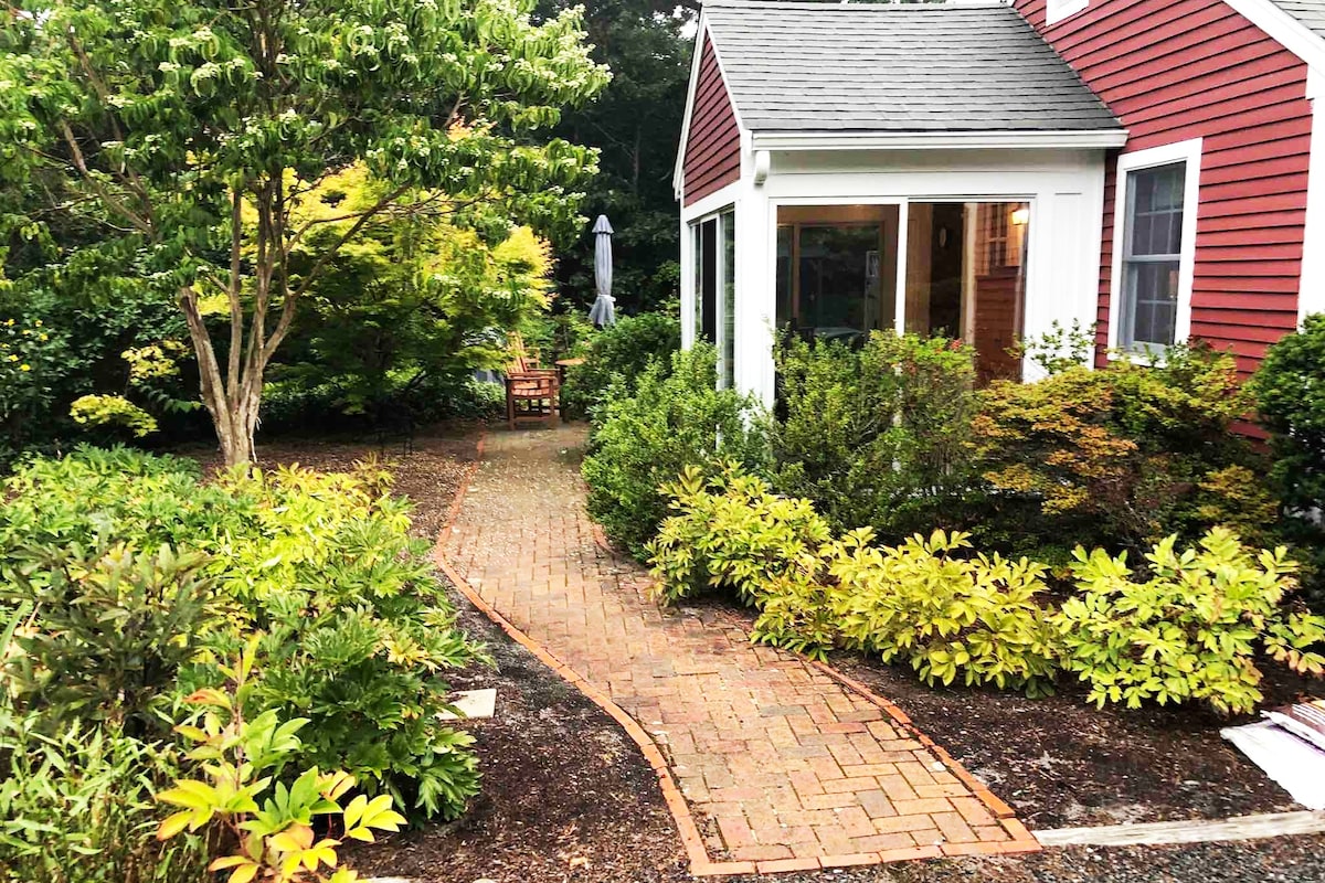 A winding brick pathway leads to the entrance of a red cottage, bordered by lush green shrubs and vibrant foliage. A small patio area with a wooden table and chairs is visible in the background, alongside a tall umbrella providing shade.