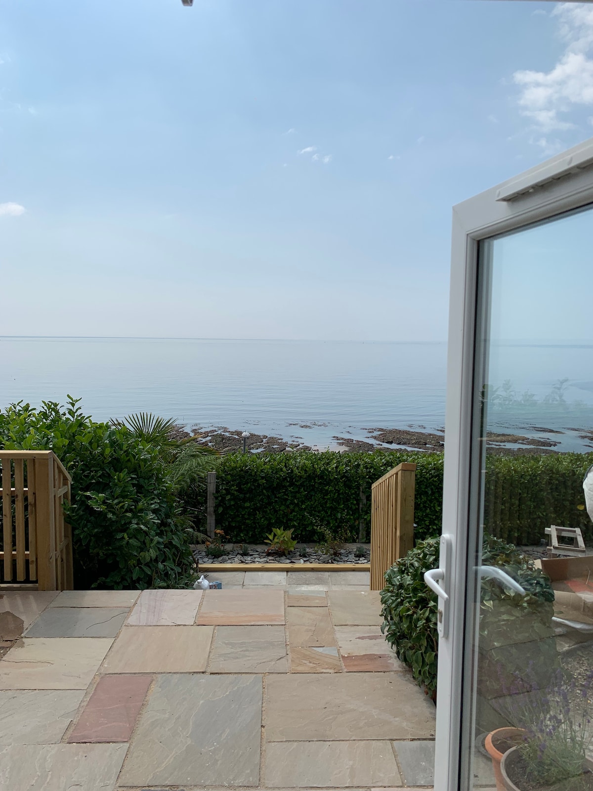 A serene view of the calm sea is framed by a glass door with a slight opening. Lush greenery and a wooden fence border the view, while a sandstone patio with scattered plants creates an inviting space for outdoor relaxation.